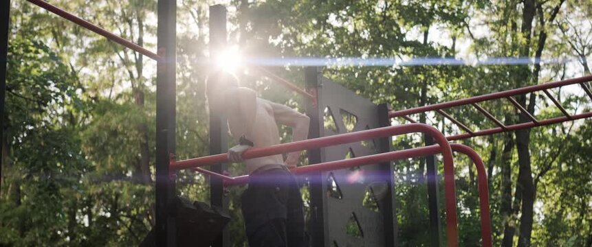 Fit shirtless athlete training chest and arms with dips on outdoor parallel bars in forest park. Handheld anamorphic shot during golden hour creates cinematic sports atmosphere.