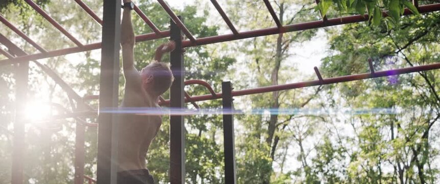 Fit male athlete walking across monkey bars during outdoor workout in green park. Handheld anamorphic shot at golden hour highlights strength, balance and cinematic sunset lighting.