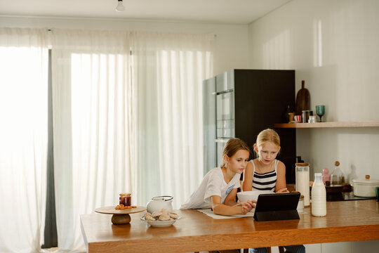 Two young girls share a tablet in a sunlit kitchen while exploring recipes. They are engaged and focused, surrounded by baking ingredients, laughter filling the air. - Powered by Adobe