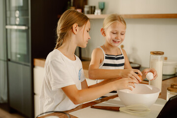 Two young girls joyfully crack eggs into a bowl while baking in a bright, modern kitchen. They share smiles as they mix ingredients, enjoying their time together.