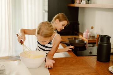 Two young girls are busy in a bright kitchen, one mixing batter energetically while the other...