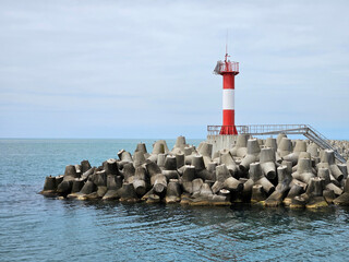 Red and white lighthouse sits on a rocky shore. The water is calm and the sky is cloudy