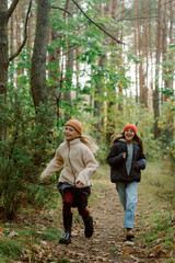 Fototapeta premium Two young girls are running excitedly down a narrow path in a vibrant forest. The trees are lush with green leaves, and the sun is shining through, creating a cheerful atmosphere.