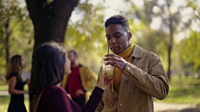 A diverse young couple shares a refreshing drink, smiling and laughing during a pleasant conversation in a sunlit park. They enjoy a relaxing moment outdoors while friends mingle in the background.