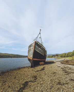 View of a weathered shipwreck rests on the stony shore under a vast sky, its dark hull contrasting with the pale beach, Corpach, Fort William, Scotland, United Kingdom.