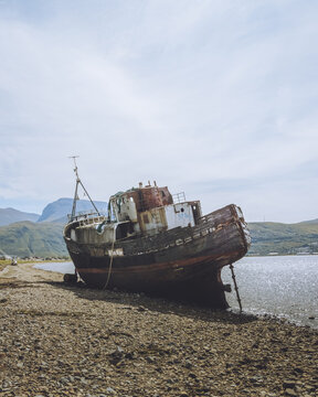 View of the rusted, decaying Corpach Shipwreck rests on a pebbled beach against the backdrop of tranquil waters and distant mountains, Fort William, Scotland, United Kingdom.