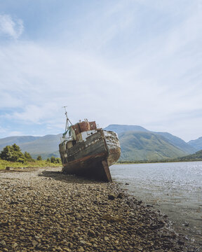 View of the weathered, decaying Corpach Shipwreck resting on the stony shore against a backdrop of majestic mountains and serene lake, Fort William, Scotland, United Kingdom.