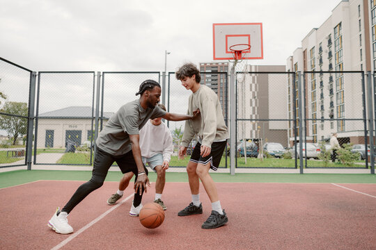 Three friends engage in a spirited basketball game on an urban court. The atmosphere is lively, with clouds overhead and buildings in the background, capturing a fun moment in the afternoon.