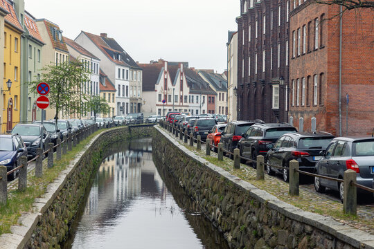 Narrow canal street in Wismar lined with parked cars on both sides beside stone embankment  water reflections. Urban oldtown scene shows full parking, historic architecture and everyday city traffic