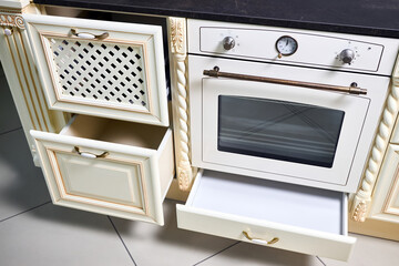 Interior of modern kitchen in classic style with golden elements in beige colors with built-in appliances. Drawer for plates or dishes shelf under the countertop with lattice front panel made of wood.