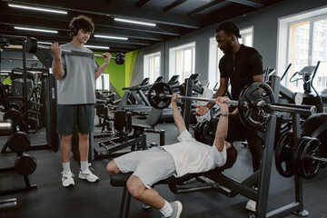 Two friends support a third as he bench presses weights in a modern gym. They share laughter and...