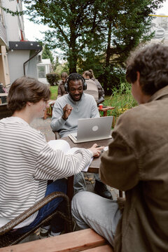 Three friends gather around a table in a lush outdoor setting, working intently on a laptop while sharing ideas and enjoying the warm sunlight of the afternoon.
