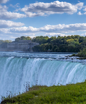View of the majestic, turquoise waterfall cascading powerfully over the precipice, framed by verdant grass and a blue sky dotted with fluffy clouds, Niagara Falls, Ontario, Canada.