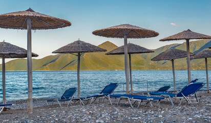 straw umbrella and sun loungers on a rocky beach in front of blue sea  and green mountain in Peloponnese Greece