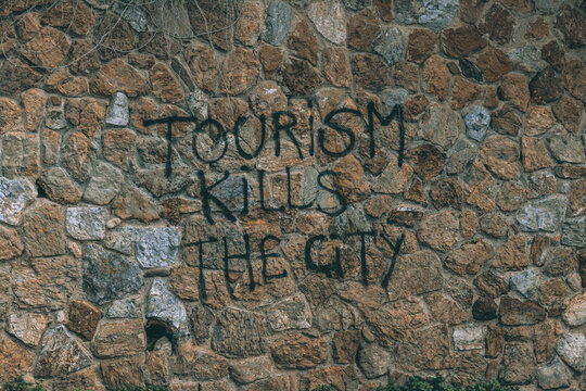 View of rough stone wall defiantly proclaims "Tourism Kills the City" in stark black graffiti, a gritty urban statement, Barcelona, Catalonia, Spain.