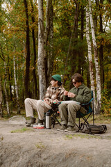 In a serene forest setting, two friends sit comfortably on folding chairs by the water, enjoying fishing together. They share laughs and good moments, surrounded by vibrant trees.