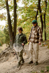 Naklejka premium Two friends walk along a forest path, preparing for a fishing adventure. They carry fishing rods and a net, surrounded by vibrant autumn leaves under the warm sun.