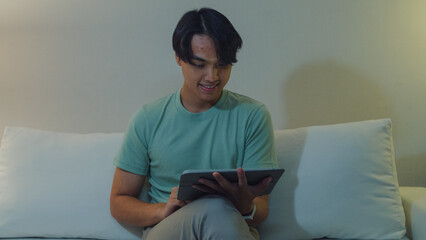 Young Asia male with casual cloth sit on sofa finger scroll digital tablet watch online movie in living room cozy house at night. E-book reading, Shopping online, Lifestyle leisure at home concept. © tirachard