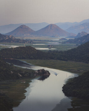 View of a serene river winding through a lush valley, framed by majestic mountains under a soft, hazy sky, Rijeka Crnojevica, Montenegro.