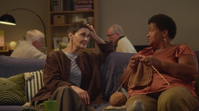 Medium long shot of senior Black woman knitting while chatting with Caucasian female friend, both spending relaxed cozy evening in nursing home with male residents playing board game in background