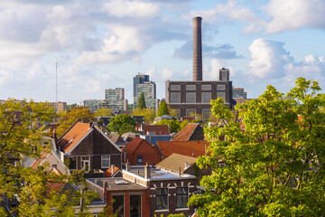 View of rooftops and vibrant green trees under a bright sky, with a tall industrial chimney and modern buildings in the distance, Leiden, South Holland, Netherlands.