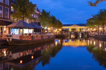 View of the serene canal reflecting the warm glow of lights from waterside restaurants and historic buildings under the twilight sky, Leiden, South Holland, Netherlands.