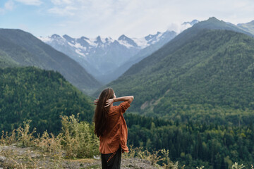 Fototapeta premium A lone traveler stands on a hillside, gazing at snow capped mountains and a vast valley, soaking in the tranquil alpine landscape and dense forest, creating a moment of awe and quiet reflection.
