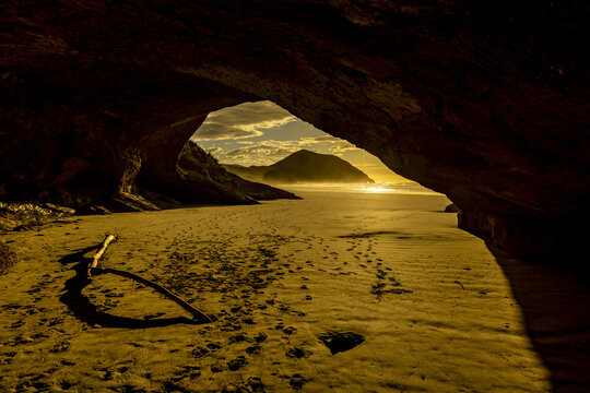 View of golden sand meeting distant mountains under a sky kissed by the setting sun, framed by the dark embrace of a cave, Pūponga, Tasman Region, New Zealand.