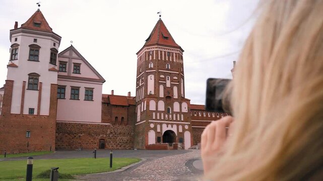 Over the shoulder view of a female tourist with blonde hair taking a picture of the historic mir castle complex in belarus with her mobile phone, capturing the beautiful medieval architecture