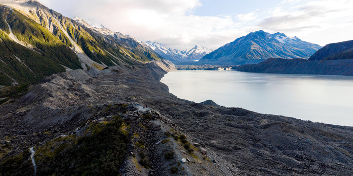Aerial view of the rugged, rocky terrain meeting the serene, glacial lake, shadowed by snow-capped mountains, Tasman Glacier, Canterbury Region, New Zealand. - Powered by Adobe