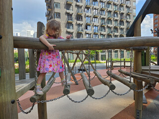 Little girl is climbing a wooden structure with chains. She is wearing a pink dress. The structure is located in a park near entrance in the courtyard of the house in residential complex
