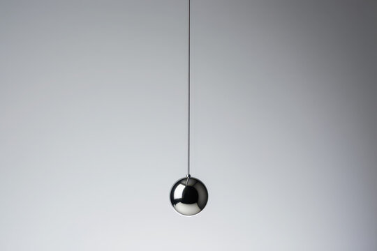 Simple chrome sphere hangs motionless, pendulum suspended against gray background. This minimal abstract concept represents physics, time, gravity, balance, and quiet focus