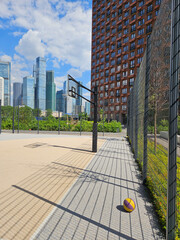Yellow and orange basketball is sitting on the ground in front of a chain link fence. The fence is located behind a basketball hoop near entrance in the courtyard of the house in residential complex