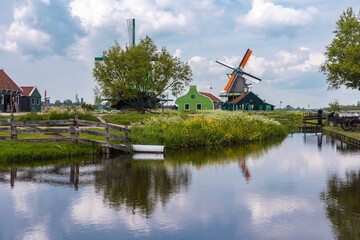 View of windmills with orange and green accents rising above a tranquil canal reflecting the cloudy sky, Zaandijk Zaanse Schans, Zaandam, Netherlands.