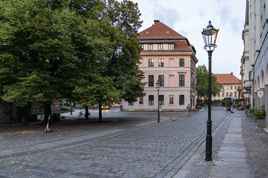 View of cobblestone streets leading to a light pink building under a canopy of green trees and glowing lamps in Nikolai Viertel, Mitte, Berlin, Germany.