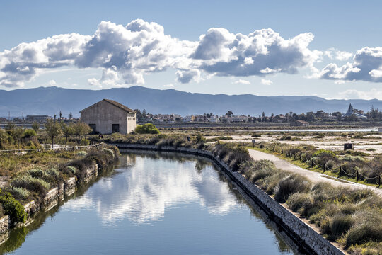 View of the tranquil canal reflecting the sky, bordered by dry grasses, leading towards a rustic building against a mountain backdrop, Cagliari, Sardinia, Italy.
