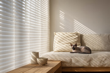 A cozy living room with soft natural light. A small gray Siamese cat rests on a beige couch. Minimalist decor includes wooden furniture and simple ceramic bowls.