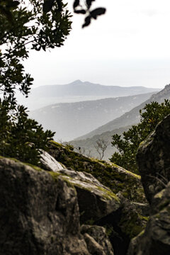 View of rugged, moss-covered rocks and verdant trees frame distant, hazy mountain peaks under a muted sky, Parco dei Sette Fratelli, Sardinia, Italy.