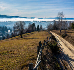 Countryside Mountain Road with Wooden Fence in Rural Landscape. Sunshine in the late autumn mountain landscape with a fence and dirt road above misty valleys, Harghita County, Livezi village, Romania.