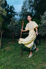 Fototapeta premium A woman in a pale yellow dress walks across a grassy field at dusk, holding a wooden stick, enjoying a peaceful outdoor moment in a natural setting.