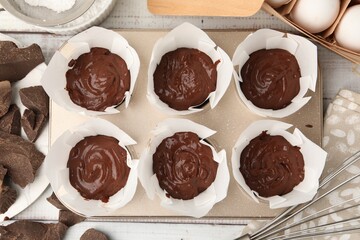 Chocolate dough in cupcake tray and ingredients for muffins on white wooden table, flat lay