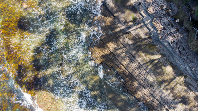 Aerial view of the rugged Tessellated Pavement kissed by the turquoise sea, its geometric patterns softened by the foamy embrace of the tide, Tessellated Pavement State Reserve, Tasmania, Australia.