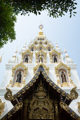 Fototapeta premium Wat Santitham, Elegant White And Gold Thai Temple Tower With Intricate Carvings, Buddha Statues, And Lush Trees