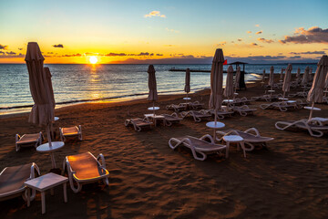 Deserted resort beach at sunset with long shadows on sand, empty sunbeds, and a golden sun path on the sea under a gradient sky. End of season calm. Mediterranean, Side, Antalya, Turkey.

