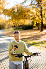Joyful Korean woman biking and texting in a beautiful autumn park under golden trees