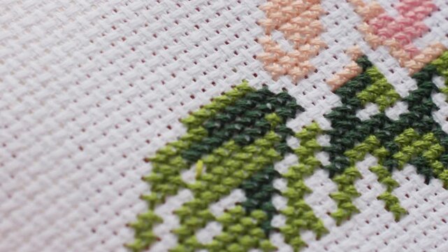 Close up designer embroidering white canvas, needle with green thread passing through cross stitch, leafy pattern forming, fine texture visible, careful craft captured under soft light