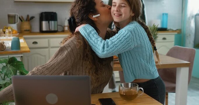 Daughter brings a cup of tea to her mother who is working online on a laptop at home. Warm family moment, care, and connection during work from home routine.