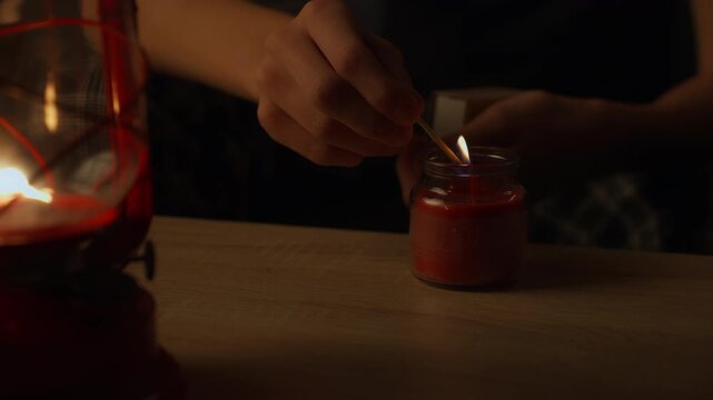 Person using matchstick to light red candle in glass jar, seeking an alternative light source during power outage, with vintage red kerosene lantern glowing in dark room