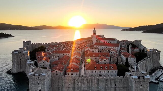 Aerial view of an ancient walled city at sunset: stone fortifications, red-roofed buildings, calm waters, sun casting golden glow