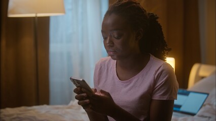 Annoyed tired upset African American woman female student girl businesswoman holding smartphone typing mobile phone mail SMS message fail working error mistake sit bed laptop at home bedroom evening